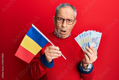 Handsome senior man with grey hair holding romania flag and leu banknotes in shock face, looking skeptical and sarcastic, surprised with open mouth