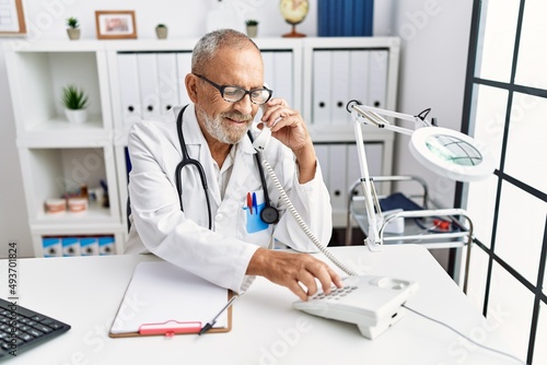 Senior grey-haired man wearing doctor uniform talking on the telephone at clinic