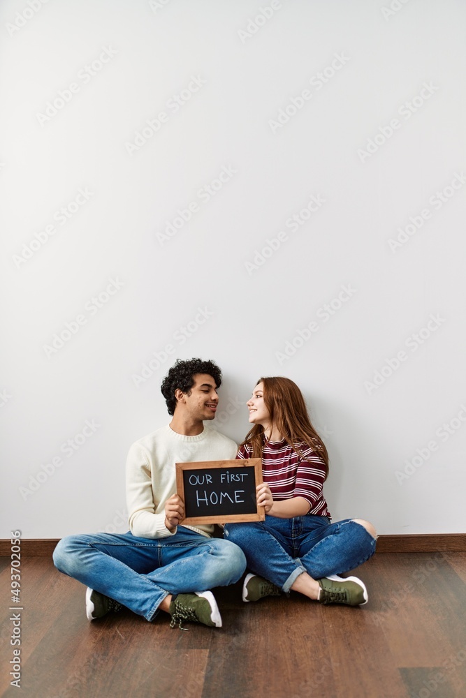 Young hispanic couple holding our first home blackboard sitting on the floor at empty new house.