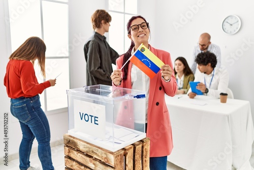 Young venezuelan voter woman smiling happy holding venezuela flag standing by ballot at vote center.