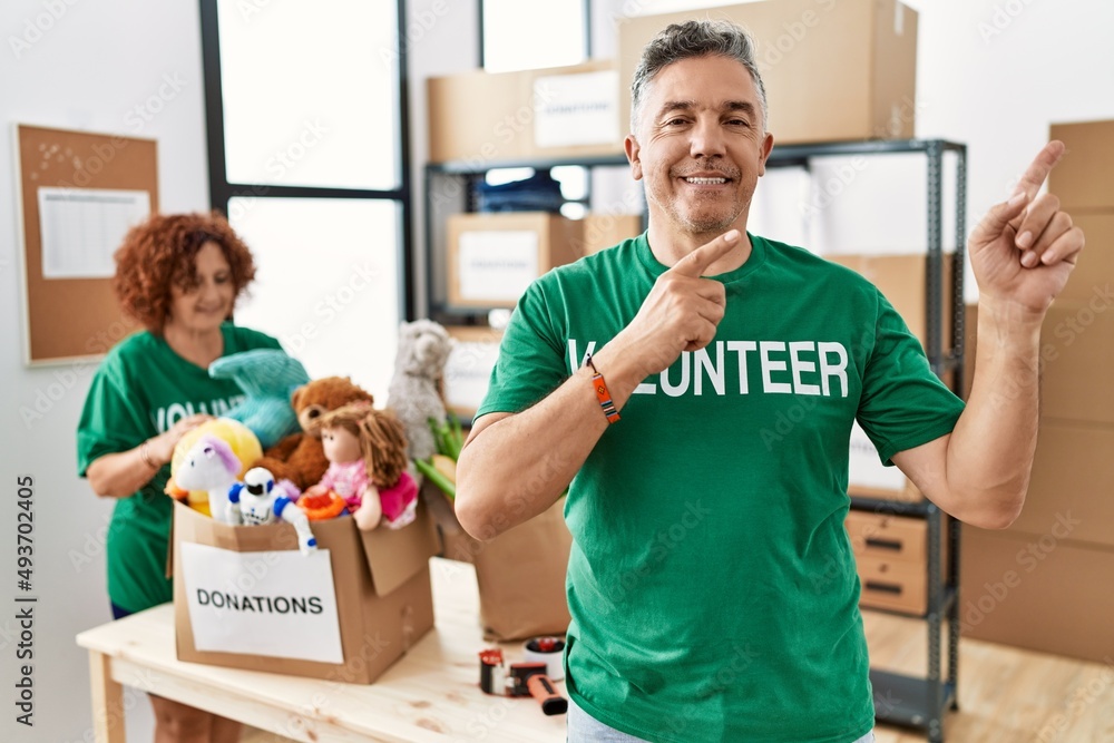 Middle age man wearing volunteer t shirt at donations stand smiling and looking at the camera pointing with two hands and fingers to the side. Middle age man wearing volunteer t shirt at donations stand smiling and looking at the camera pointing with two hands and fingers to the side.