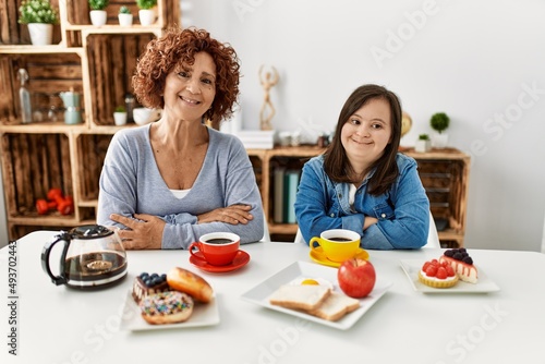 Family of mother and down syndrome daughter sitting at home eating breakfast happy face smiling with crossed arms looking at the camera. positive person.