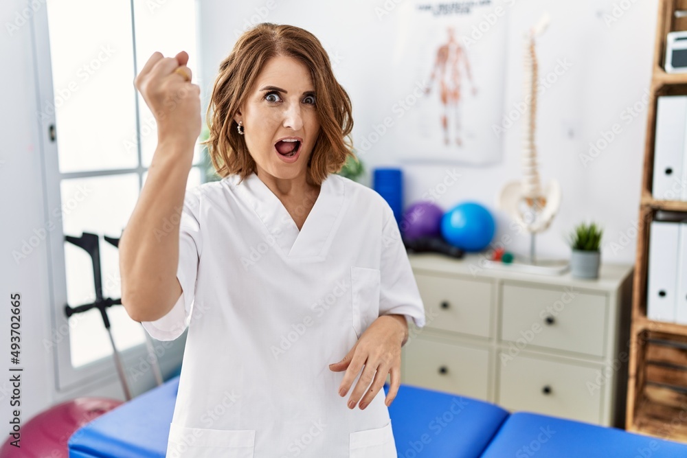 Middle age physiotherapist woman working at pain recovery clinic angry and mad raising fist frustrated and furious while shouting with anger. rage and aggressive concept. Middle age physiotherapist woman working at pain recovery clinic angry and mad raising fist frustrated and furious while shouting with anger. rage and aggressive concept.