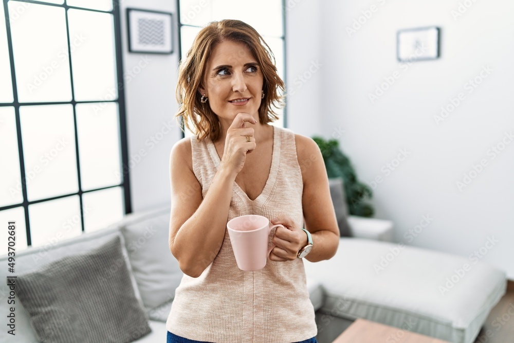 Middle age woman drinking a cup coffee at home with hand on chin thinking about question, pensive expression. smiling and thoughtful face. doubt concept. Middle age woman drinking a cup coffee at home with hand on chin thinking about question, pensive expression. smiling and thoughtful face. doubt concept.