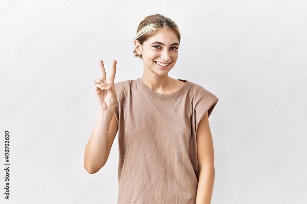 Young blonde woman standing over isolated background smiling with happy face winking at the camera doing victory sign. number two. Young blonde woman standing over isolated background smiling with happy face winking at the camera doing victory sign. number two.