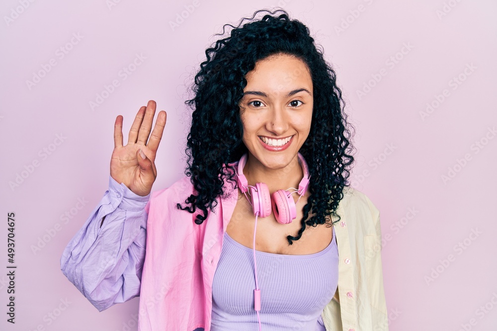 Young hispanic woman with curly hair wearing gym clothes and using headphones showing and pointing up with fingers number four while smiling confident and happy. Young hispanic woman with curly hair wearing gym clothes and using headphones showing and pointing up with fingers number four while smiling confident and happy.