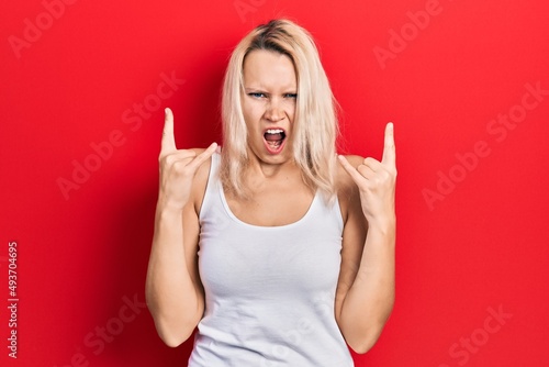 Beautiful caucasian blonde woman wearing casual white t shirt shouting with crazy expression doing rock symbol with hands up. music star. heavy concept.