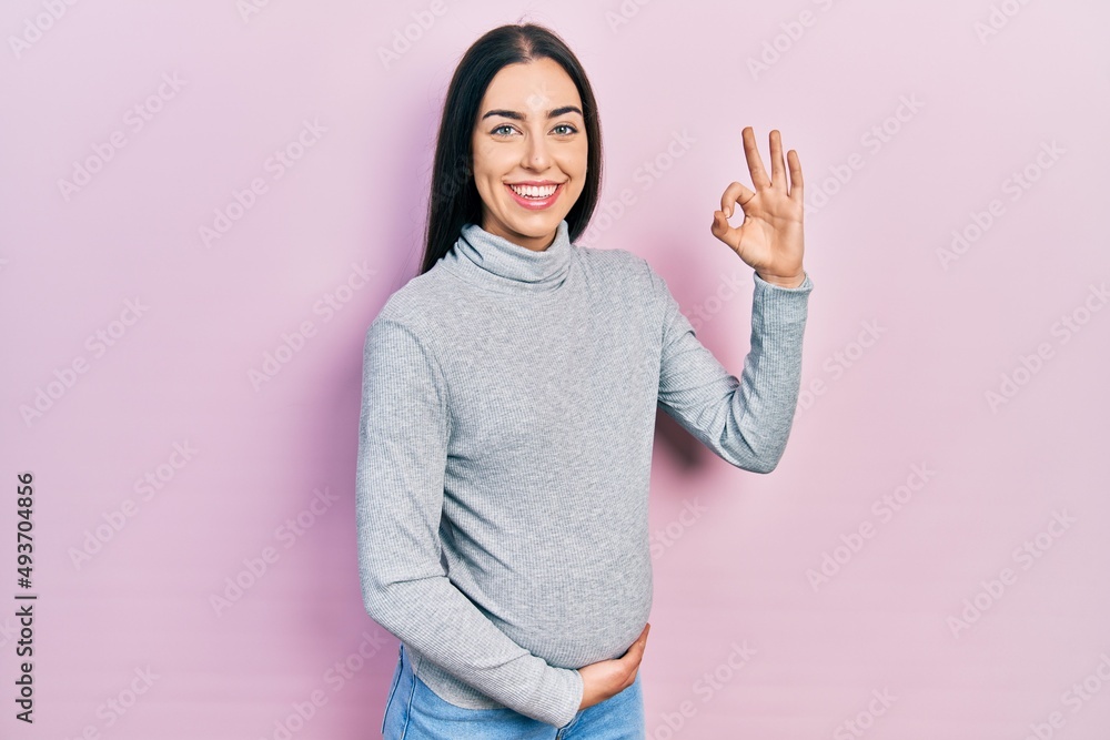 Beautiful woman with blue eyes expecting a baby, touching pregnant belly smiling positive doing ok sign with hand and fingers. successful expression. Beautiful woman with blue eyes expecting a baby, touching pregnant belly smiling positive doing ok sign with hand and fingers. successful expression.