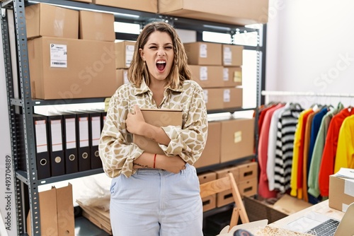 Young hispanic shopkeeper woman holding cardboard box at storeware angry and mad screaming frustrated and furious, shouting with anger. rage and aggressive concept. photo