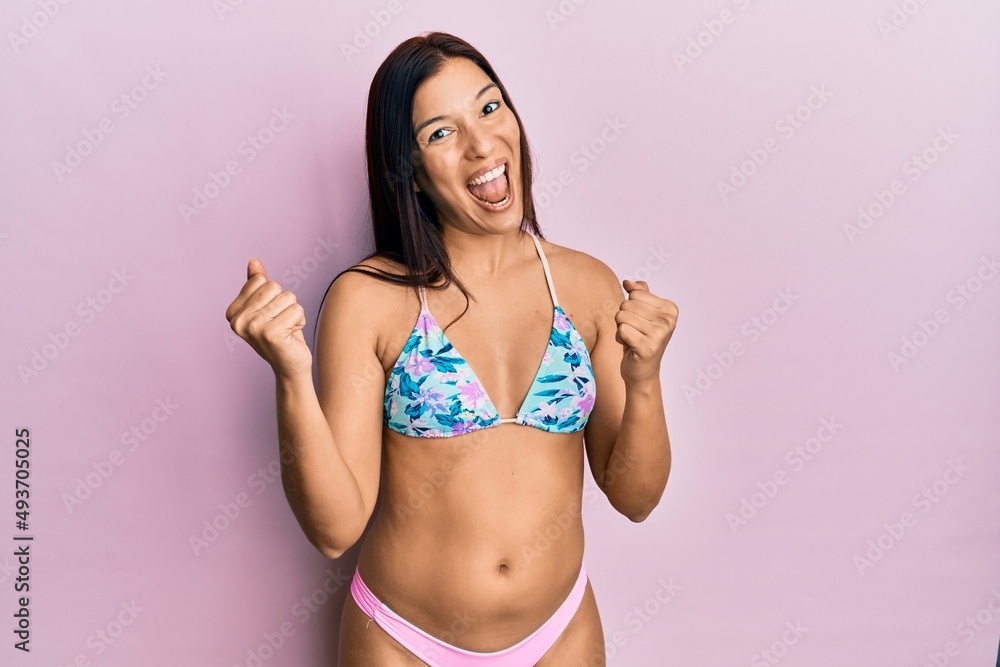 Young latin woman wearing bikini screaming proud, celebrating victory and success very excited with raised arms