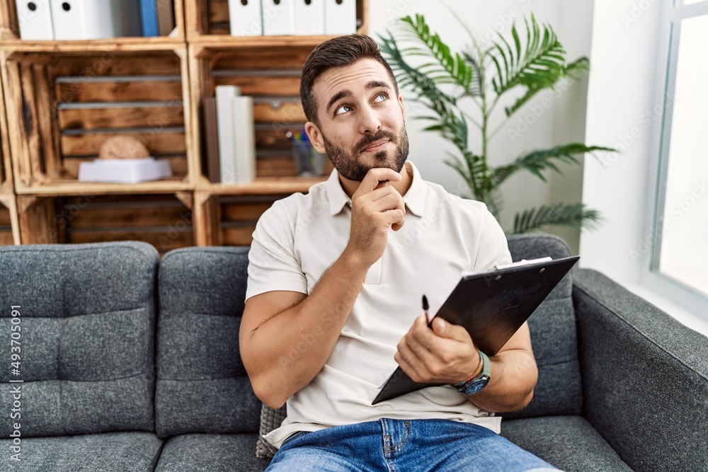 Handsome hispanic man holding clipboard working at psychology clinic with hand on chin thinking about question, pensive expression. smiling and thoughtful face. doubt concept. Handsome hispanic man holding clipboard working at psychology clinic with hand on chin thinking about question, pensive expression. smiling and thoughtful face. doubt concept.