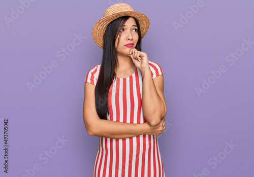 Young hispanic woman wearing summer hat serious face thinking about question with hand on chin, thoughtful about confusing idea