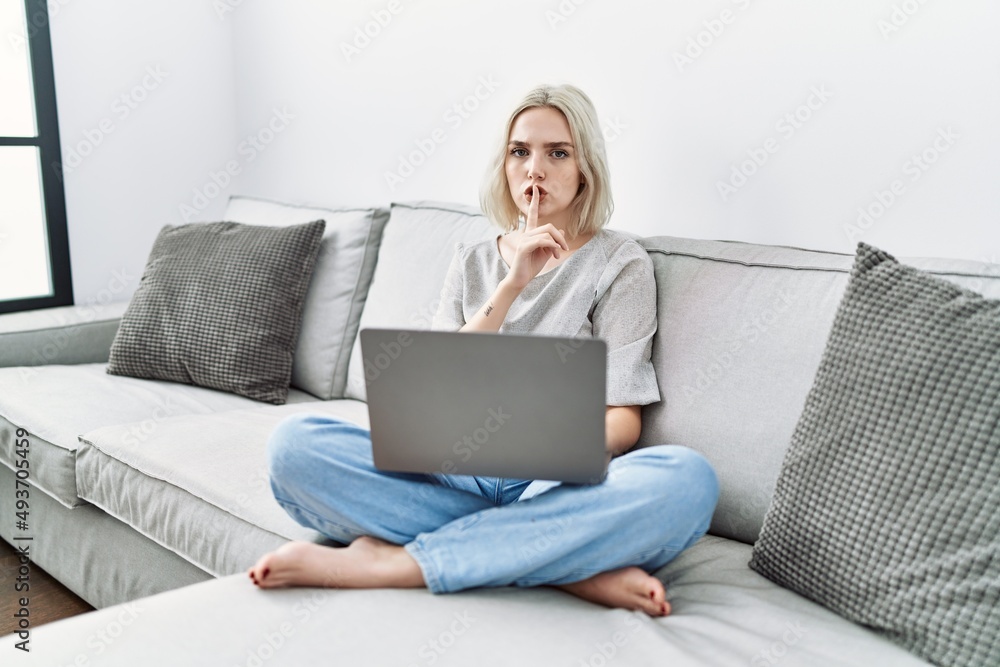 Young caucasian woman using laptop at home sitting on the sofa asking to be quiet with finger on lips. silence and secret concept. Young caucasian woman using laptop at home sitting on the sofa asking to be quiet with finger on lips. silence and secret concept.