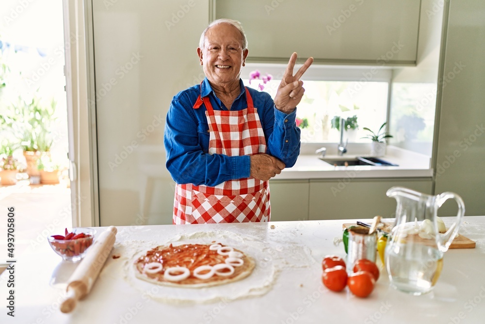 Senior man with grey hair cooking pizza at home kitchen smiling with happy face winking at the camera doing victory sign. number two. Senior man with grey hair cooking pizza at home kitchen smiling with happy face winking at the camera doing victory sign. number two.