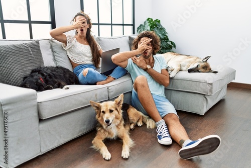 Photography Young hispanic couple with dogs relaxing at home peeking in shock covering face and eyes with hand, looking through fingers with embarrassed expression
