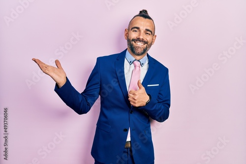 Young hispanic man wearing business suit and tie showing palm hand and doing ok gesture with thumbs up, smiling happy and cheerful