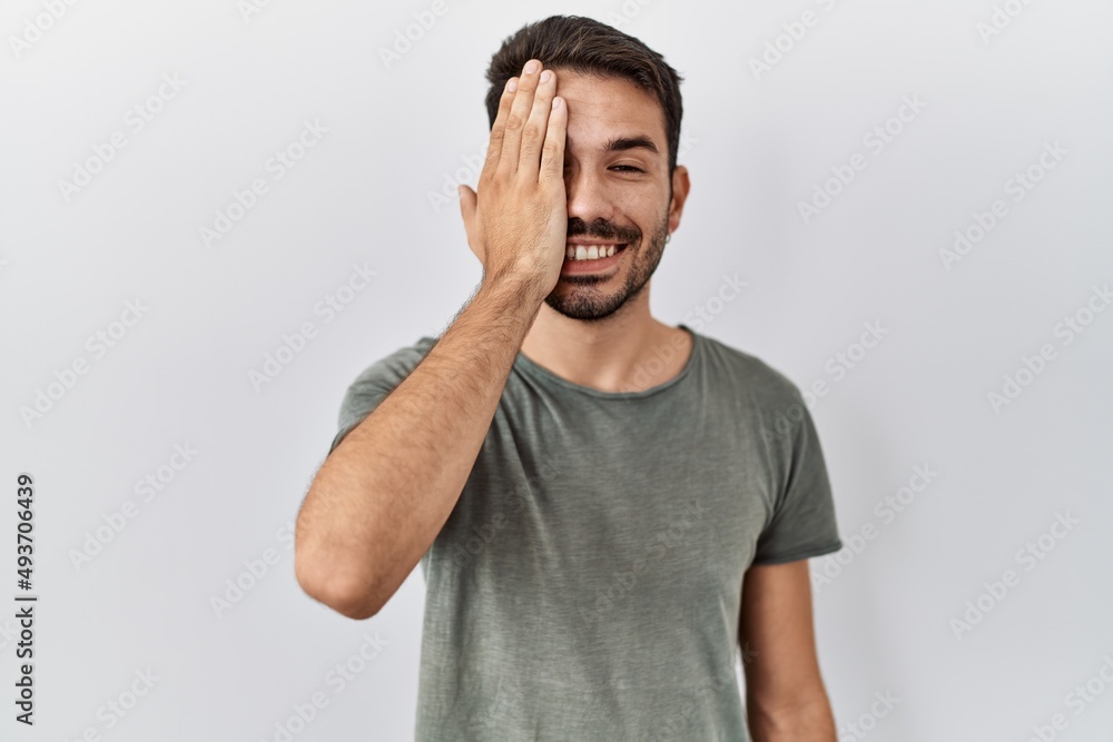 Young hispanic man with beard wearing casual t shirt over white background covering one eye with hand, confident smile on face and surprise emotion. Young hispanic man with beard wearing casual t shirt over white background covering one eye with hand, confident smile on face and surprise emotion.