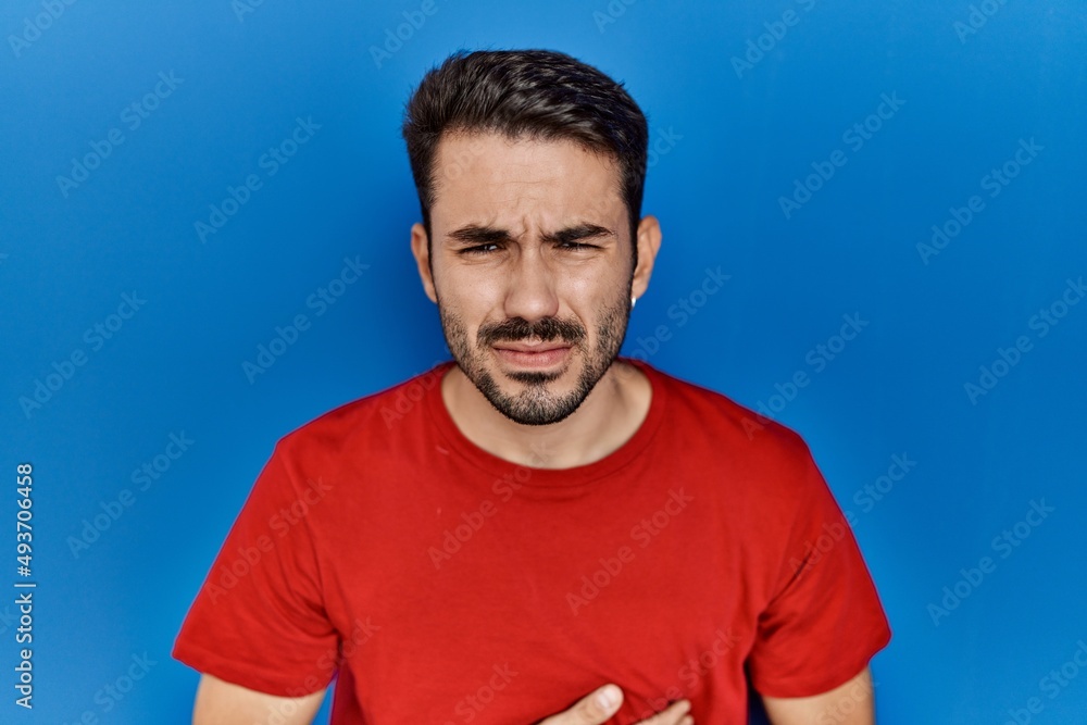 Fototapeta premium Young hispanic man with beard wearing red t shirt over blue background with hand on stomach because indigestion, painful illness feeling unwell. ache concept.