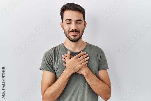 Young hispanic man with beard wearing casual t shirt over white background smiling with hands on chest with closed eyes and grateful gesture on face. health concept.