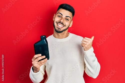 Young hispanic man with beard holding motor oil bottle pointing thumb up to the side smiling happy with open mouth