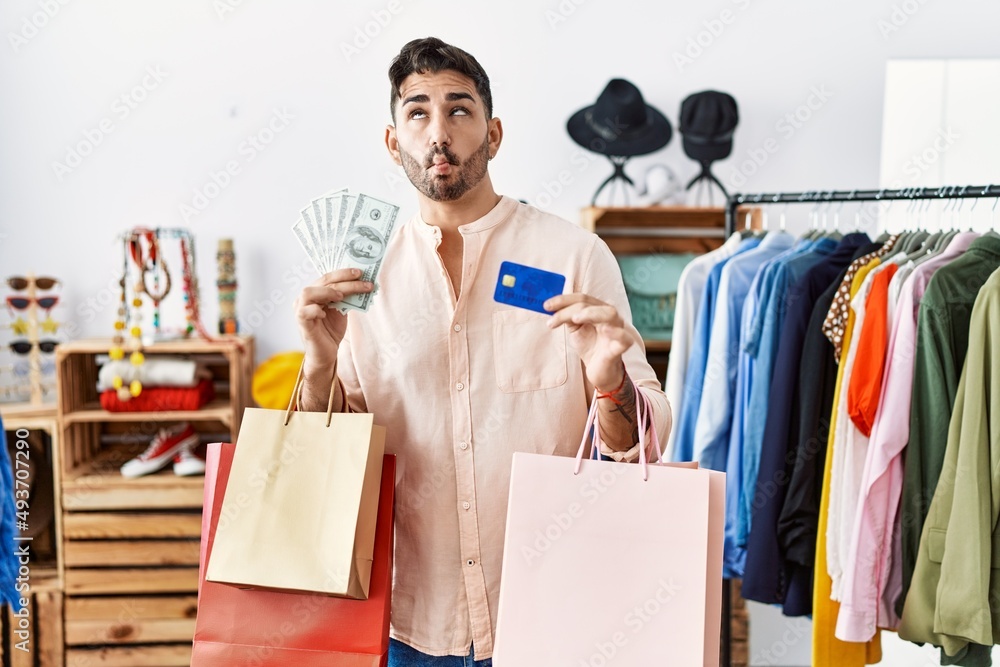 Young hispanic man holding shopping bags and credit card making fish face with mouth and squinting eyes, crazy and comical. Young hispanic man holding shopping bags and credit card making fish face with mouth and squinting eyes, crazy and comical.
