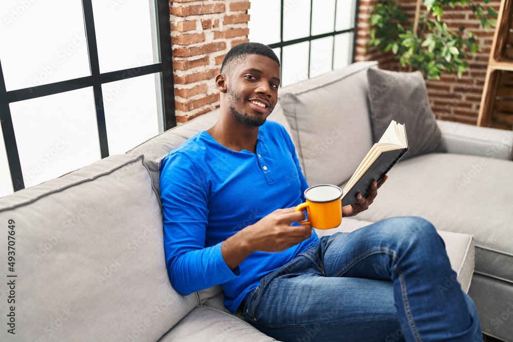 Young african american man reading book and drinking coffee sitting on sofa at home