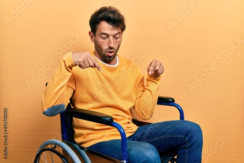 Handsome man with beard sitting on wheelchair pointing down with fingers showing advertisement, surprised face and open mouth