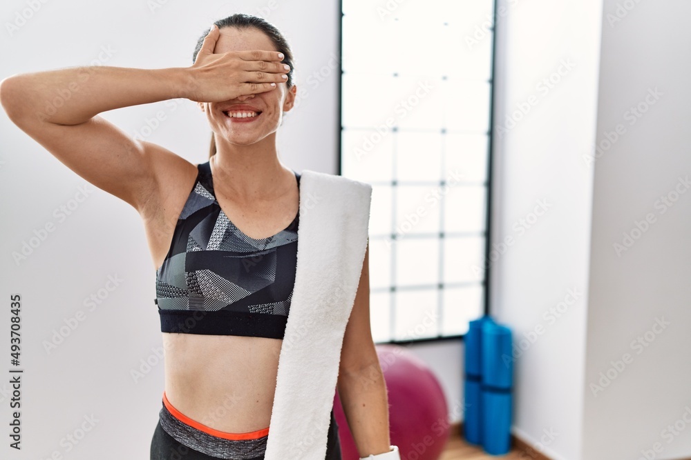 Young brunette woman wearing sportswear and towel at the gym smiling and laughing with hand on face covering eyes for surprise. blind concept.