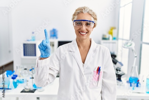 Middle age blonde woman working at scientist laboratory showing and pointing up with finger number one while smiling confident and happy.