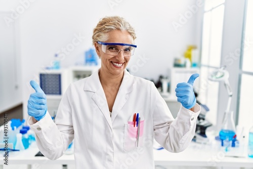 Middle age blonde woman working at scientist laboratory success sign doing positive gesture with hand, thumbs up smiling and happy. cheerful expression and winner gesture.