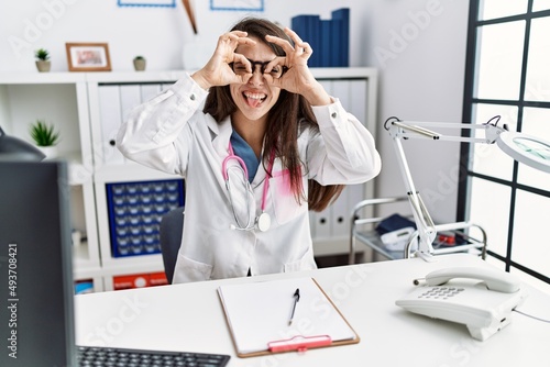 Young doctor woman wearing doctor uniform and stethoscope at the clinic doing ok gesture like binoculars sticking tongue out  eyes looking through fingers. crazy expression.