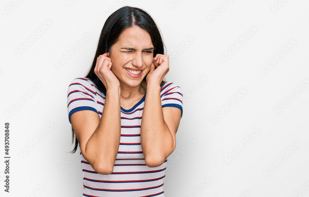 Young hispanic girl wearing casual striped t shirt covering ears with fingers with annoyed expression for the noise of loud music. deaf concept. Young hispanic girl wearing casual striped t shirt covering ears with fingers with annoyed expression for the noise of loud music. deaf concept.