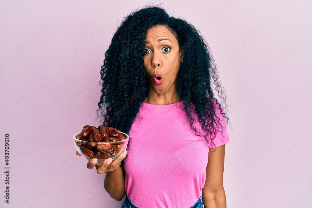 Middle age african american woman holding bowl with dates scared and amazed with open mouth for surprise, disbelief face Middle age african american woman holding bowl with dates scared and amazed with open mouth for surprise, disbelief face