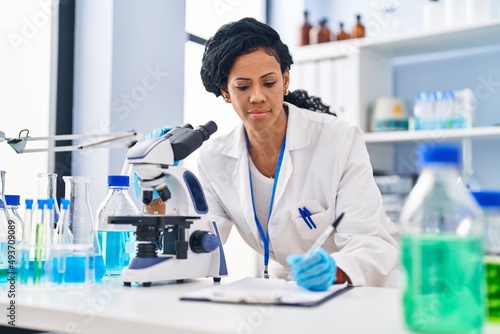 African american woman wearing scientist uniform using microscope write on document at laboratory