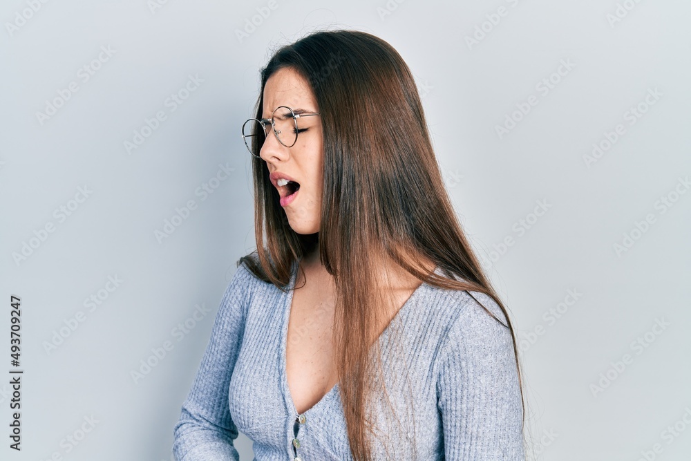 Young brunette teenager wearing casual sweater and glasses with hand on stomach because nausea, painful disease feeling unwell. ache concept. Young brunette teenager wearing casual sweater and glasses with hand on stomach because nausea, painful disease feeling unwell. ache concept.