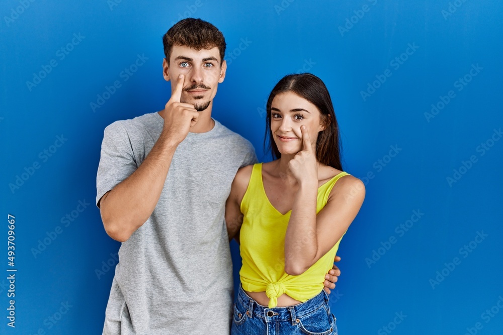 Fototapeta premium Young hispanic couple standing together over blue background pointing to the eye watching you gesture, suspicious expression
