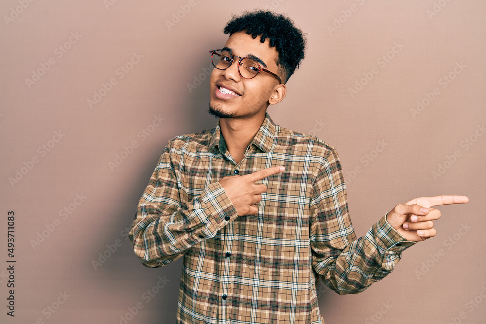 Young african american man wearing casual clothes and glasses smiling and looking at the camera pointing with two hands and fingers to the side. Young african american man wearing casual clothes and glasses smiling and looking at the camera pointing with two hands and fingers to the side.