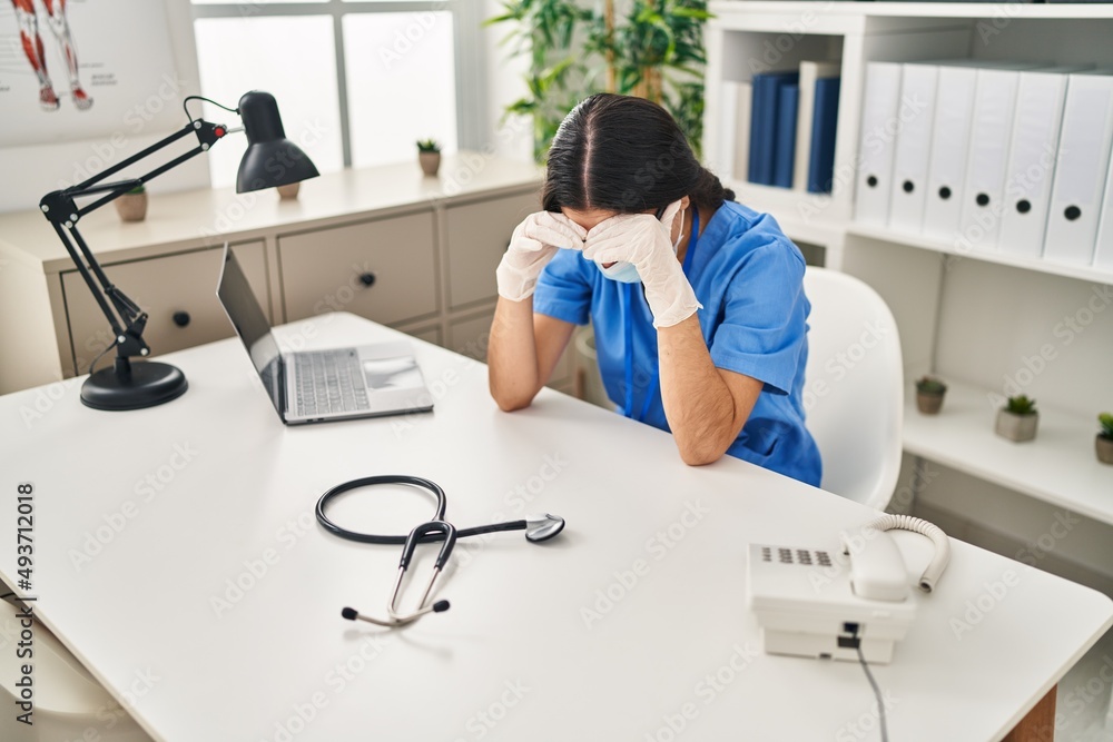 Obraz premium Young hispanic woman wearing doctor uniform and medical mask exhausted at clinic