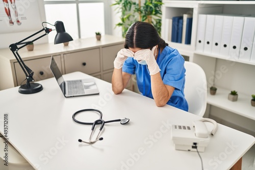 Young hispanic woman wearing doctor uniform and medical mask exhausted at clinic
