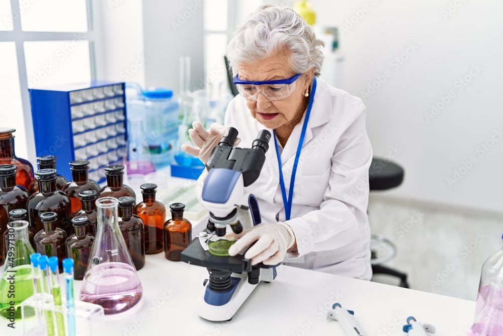 Senior grey-haired woman wearing scientist uniform using microscope at laboratory Senior grey-haired woman wearing scientist uniform using microscope at laboratory