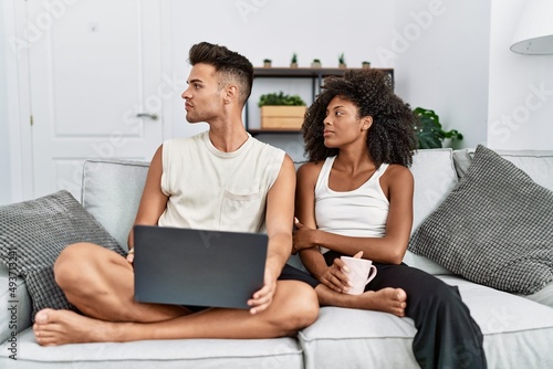 Young interracial couple using laptop at home sitting on the sofa looking to side, relax profile pose with natural face and confident smile.