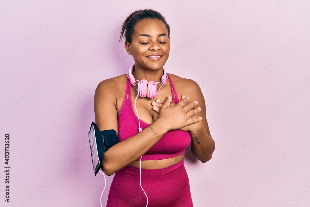 Young african american girl wearing gym clothes and using headphones smiling with hands on chest with closed eyes and grateful gesture on face. health concept.