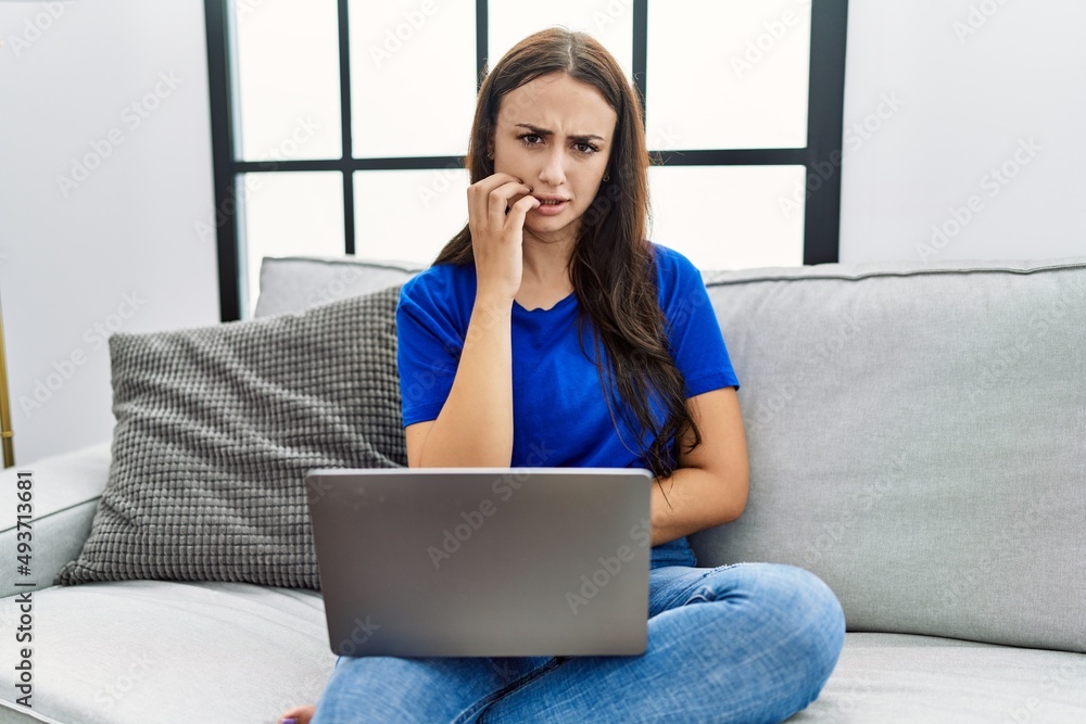 Naklejka premium Young brunette woman using laptop at home looking stressed and nervous with hands on mouth biting nails. anxiety problem.