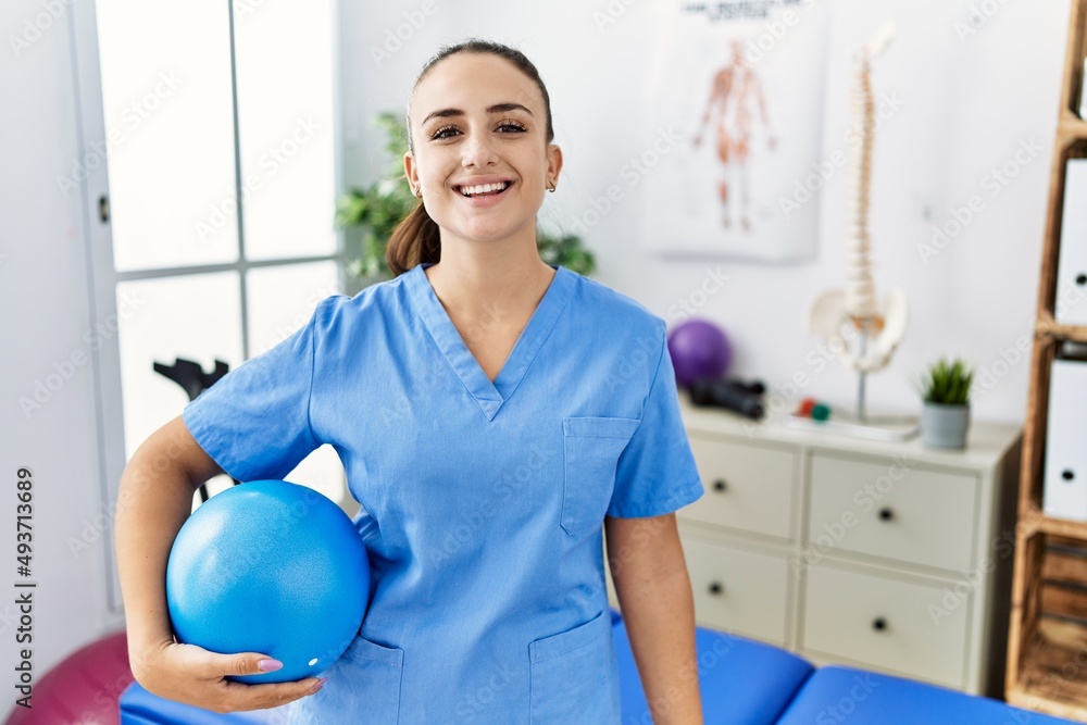 Young physiotherapist woman holding pilates ball at medical clinic looking positive and happy standing and smiling with a confident smile showing teeth Young physiotherapist woman holding pilates ball at medical clinic looking positive and happy standing and smiling with a confident smile showing teeth