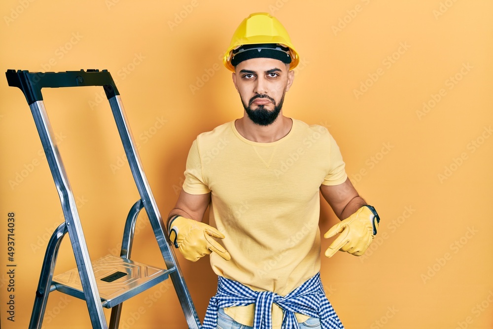 Handsome man with beard by construction stairs wearing hardhat pointing down looking sad and upset, indicating direction with fingers, unhappy and depressed. Handsome man with beard by construction stairs wearing hardhat pointing down looking sad and upset, indicating direction with fingers, unhappy and depressed.