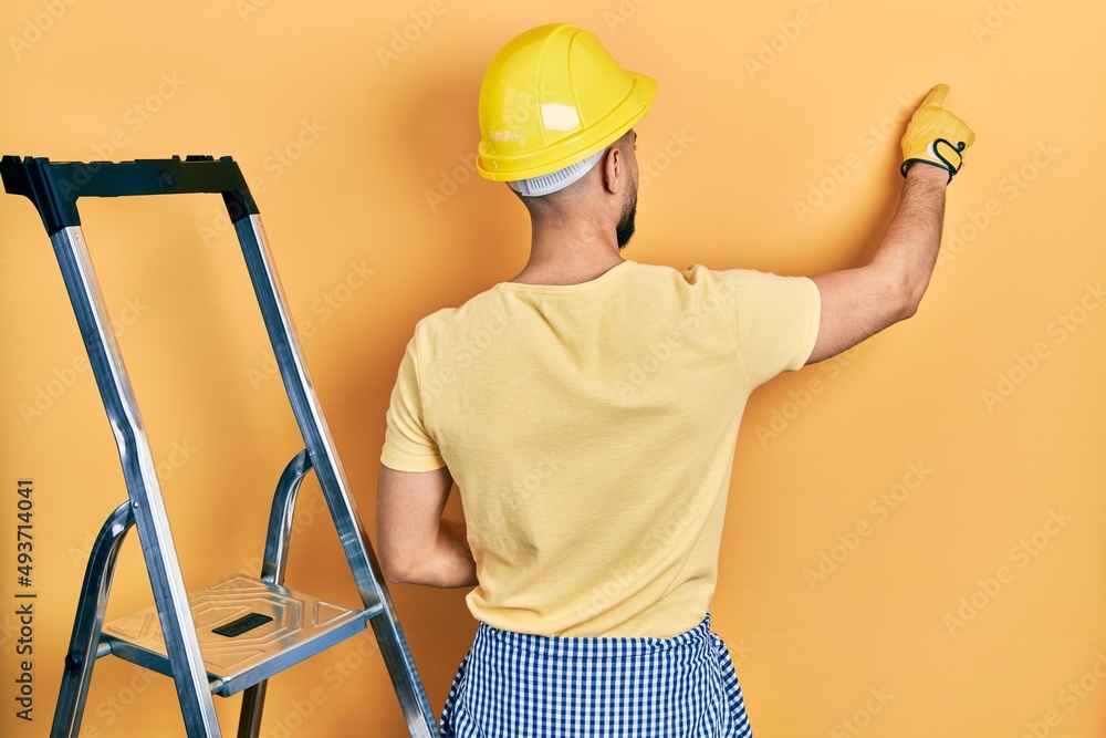 Handsome man with beard by construction stairs wearing hardhat posing backwards pointing ahead with finger hand Handsome man with beard by construction stairs wearing hardhat posing backwards pointing ahead with finger hand