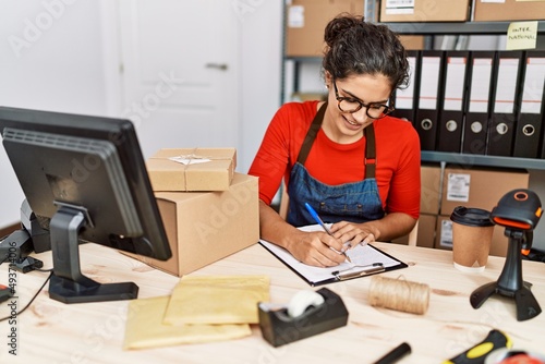 Young latin woman ecommerce business worker writing on clipboard at office