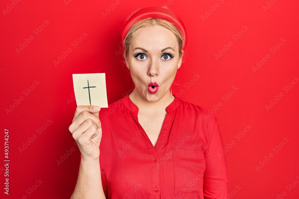 Young blonde woman holding catholic cross reminder scared and amazed with open mouth for surprise, disbelief face Young blonde woman holding catholic cross reminder scared and amazed with open mouth for surprise, disbelief face