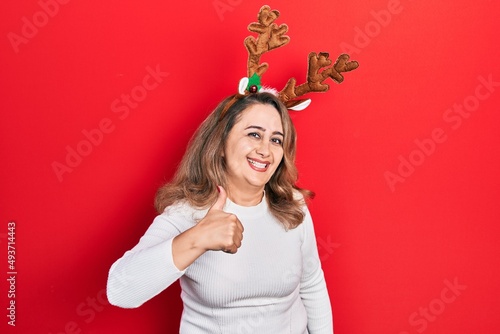 Middle age caucasian woman wearing cute christmas reindeer horns doing happy thumbs up gesture with hand. approving expression looking at the camera showing success.