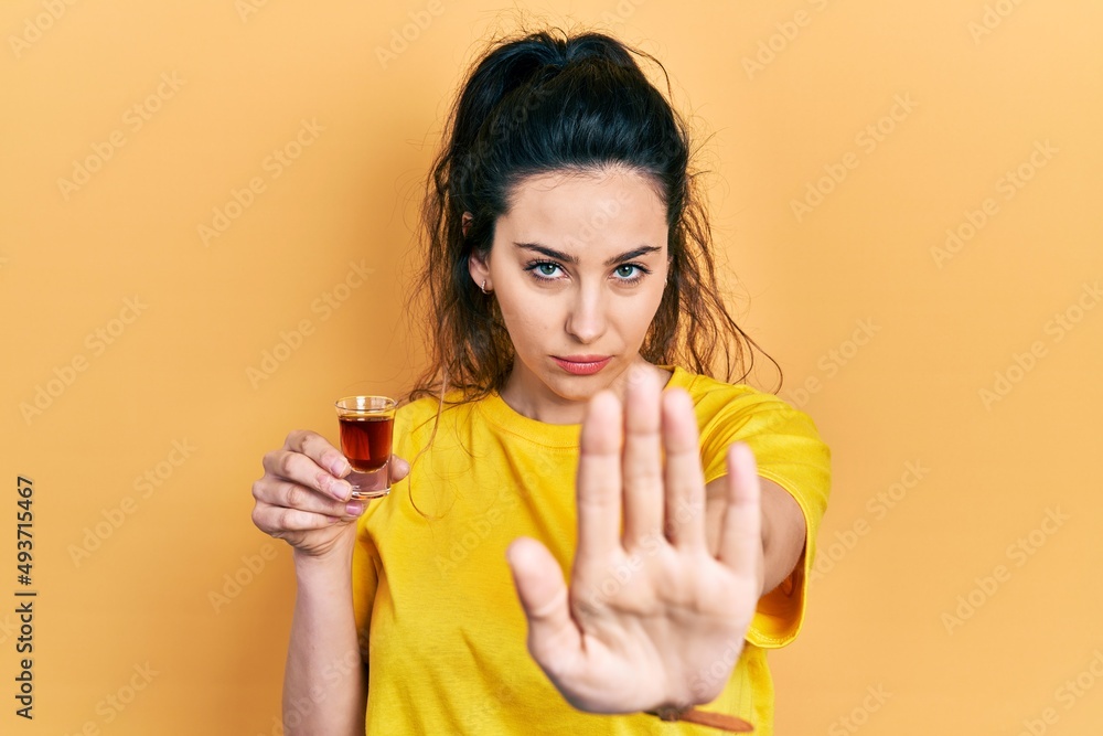 Young hispanic woman drinking whiskey shot with open hand doing stop sign with serious and confident expression, defense gesture Young hispanic woman drinking whiskey shot with open hand doing stop sign with serious and confident expression, defense gesture